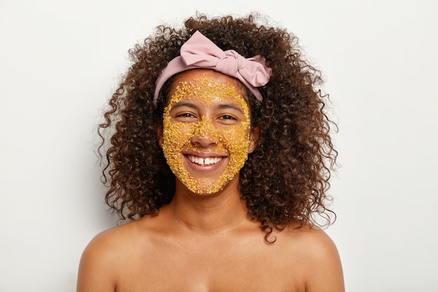 陽気なアフリカ系アメリカ人女性のヘッドショットは、顔に自然なスクラブマスクを適用し、輝きを追加し、死んだ皮膚をスクラブし、さまざまな細菌を殺します。ヘッドバンドは縮れた髪の裸の肩を持っています。美容コンセプト