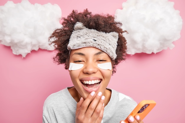 Headshot of cheerful curly haired woman smiles broadly laughs at something dressed in home wear uses modern cellphone for surfing social network