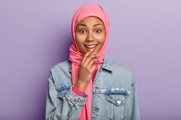 밝고 어두운 피부를 가진 무슬림 여성의 얼굴 사진은 부드러운 미소를 지으며 하얀 치아를 보여주고 분홍색 히잡을 입습니다.