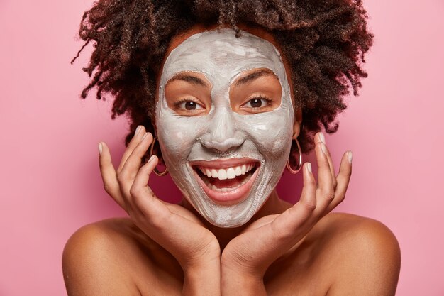 Headshot of cheerful girl with white clay mask, touches face, takes care of skin and beauty, has positive smile, Afro haircut, models over pink wall, poses indoor. Facial treatment concept