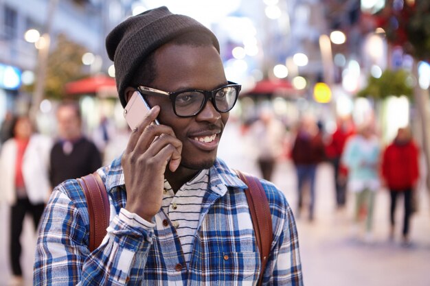 Headshot of cheerful modern young African tourist with backpack wearing hat and glasses having phone conversation while walking along crowded street during summer vacations in foreign country