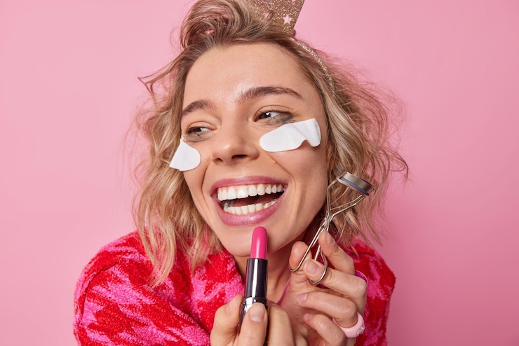 Free Photo headshot of cheerful woman applies lipstick does makeup holds eyelashes curler smiles broadly undergoes beauty procedures has leaked makeup hairstyle focused away poss against pink studio wall