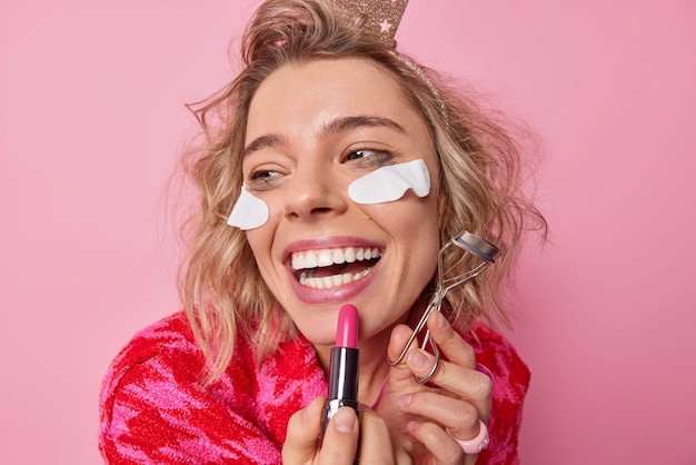 Headshot of cheerful woman applies lipstick does makeup holds eyelashes curler smiles broadly undergoes beauty procedures has leaked makeup hairstyle focused away poss against pink studio wall