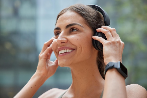 Headshot of cheerful woman puts on wireless headphones listens favorite audio track wears smartwatch on wrist has happy expression strolls outdoors against blurred background. Lifestyle and hobby
