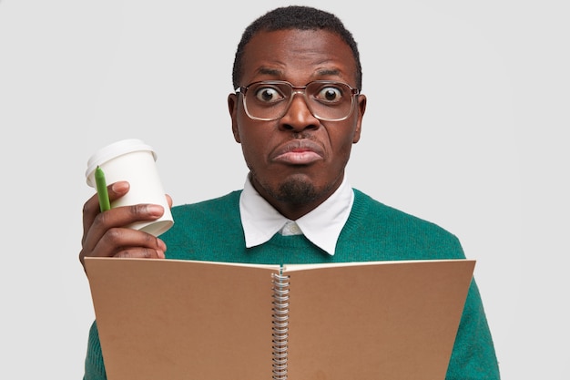 Headshot of confused black male student wonk looks in bewilderment, writes down text information at notebook