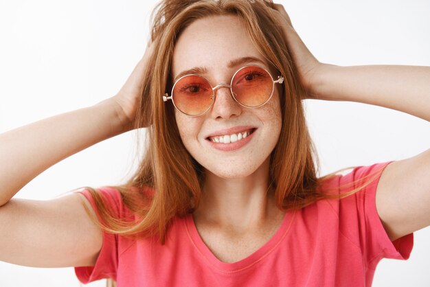 Headshot of creative and happy attractive ginger girl with cute freckles in stylish pink sunglasses touching hairstyle and smiling broadly enjoying new look while watching in mirror satisfied, pleased