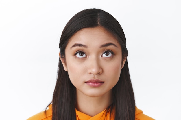 Headshot of curious cute asian girl with short dark hair looking up, observe something interesting upwards, thinking or contemplating intriguing scene, standing white wall