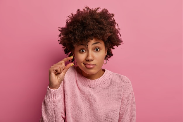 Il primo piano del volto della donna dai capelli ricci discute di qualcosa di molto piccolo, modella qualcosa di molto piccolo, stringe le labbra, vestito con un maglione casual, isolato sul muro rosa, chiede un oggetto minuscolo, fa un piccolo gesto