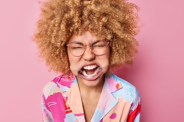 Headshot of curly haired woman with expander in mouth shows white teeth keeps eyes closed wears spectacles and colorful shirt poses against pink background Emotional female uses dental retractor