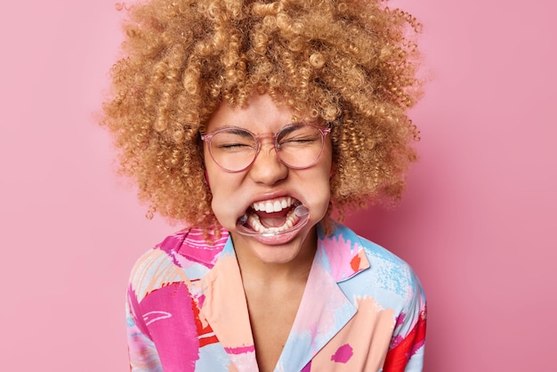 Headshot of curly haired woman with expander in mouth shows white teeth keeps eyes closed wears spectacles and colorful shirt poses against pink background Emotional female uses dental retractor