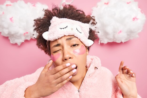 Headshot of curly haired young woman covers mouth with hand has sleepy expression wakes up early in morning undergoes beauty treatments after awakening wears comfortable nightwear