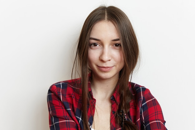 Headshot of cute European woman in love can't stop smiling, looking with happy joyful expression on her face. Beautiful young woman dressed in casual checkered shirt having rest indoors