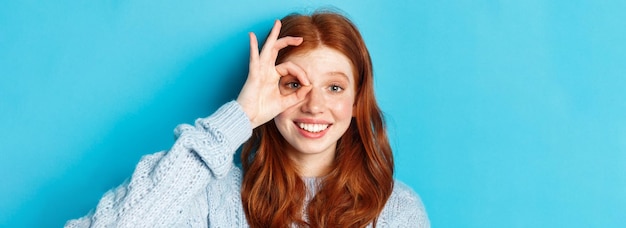 Free photo headshot of cute teenage redhead girl showing okay sign on eye and smiling standing happy against bl