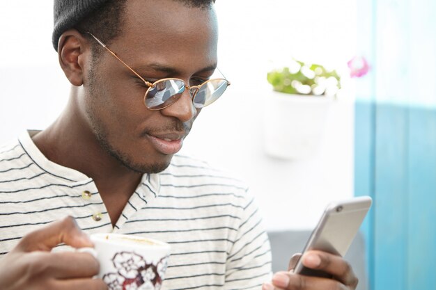 Headshot of dark-skinned freelancer drinking coffee or tea sitting in open-air restaurant or cafeteria, wearing fashionable mirrored sunglasses