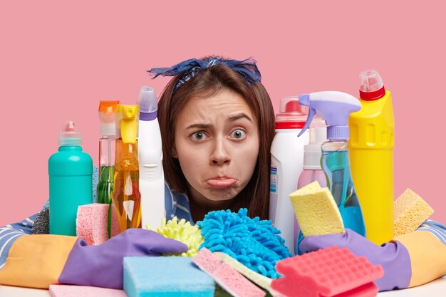 Headshot of discontent young woman purses lower lip with dissatisfaction, has much work, embraces many chemical detergents and sponges