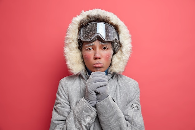 Headshot of displeased woman with cold face keeps hands together looks with imploring expression, wears warm winter jacke