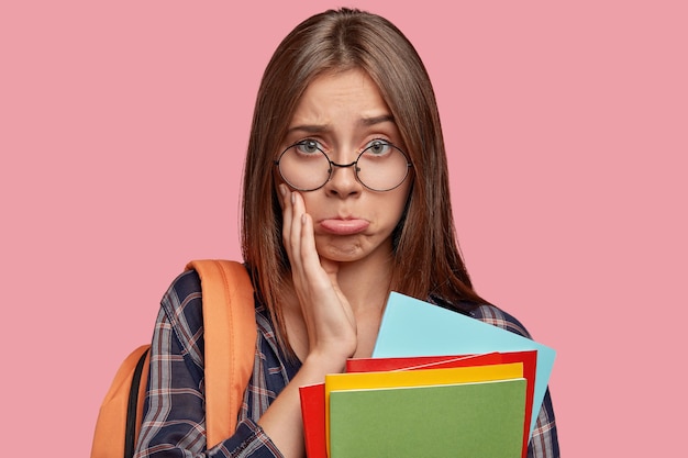 Headshot of dissatisfied schoolgirl purses lips with offensive look, has sad expression, wears round spectacles, carries rucksack