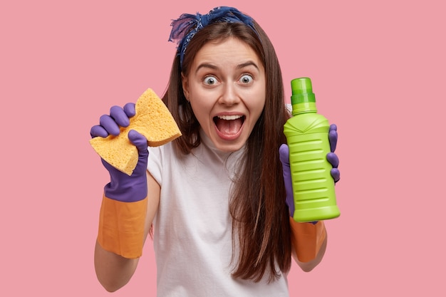 Foto gratuita primo piano del volto emotivo di una donna dai capelli scuri con gli occhi infastiditi, indossa la fascia, porta una spugna e una bottiglia di detersivo