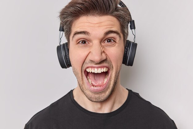 Headshot of emotional man with bristle screams loudly keeps mouth opened feels outraged listens music via wireless headphones dressed casually isolated over white background yells at someone.