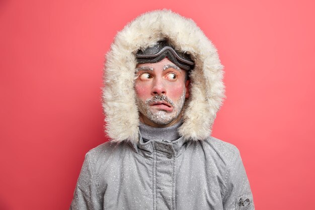 Headshot of emotive puzzled man purses lips and looks aside trembles from low temperature needs to warm wears grey jacket with fur hood.