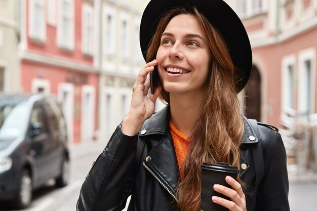 Headshot of female passenger makes phone conversation, looks happily somewhere, stands against blurred urban space