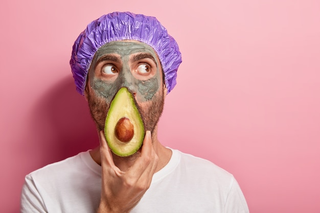 Headshot of focused man looks up, keeps slice of avocado near face, applies clay mask, removes black dots on skin