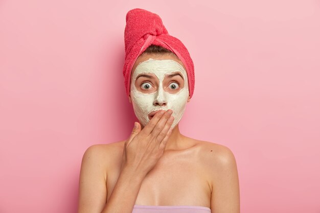 Headshot of frightened woman stares with bugged eyes, covers mouth in omg expression, wrapped towel on head, applies clay mask on face, improves skin