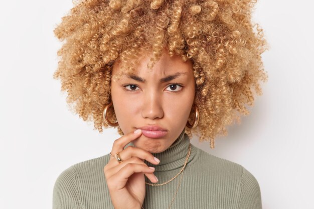 Headshot of frustrated unhappy woman purses lips looks displeased at camera feels sad or annoyed wears casual turtleneck isolated over white background thinks about something focused forward