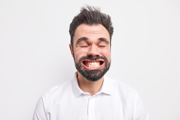 Headshot of funny bearded adult European man clenches teeth wears shirt closes eyes makes funny grimace dressed in shirt 