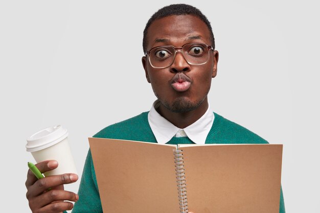 Смешной студент-мужчина надувает губы, гримасничает, носит большие оптические очки, строгую белую рубашку под свитером, держит кофе на вынос