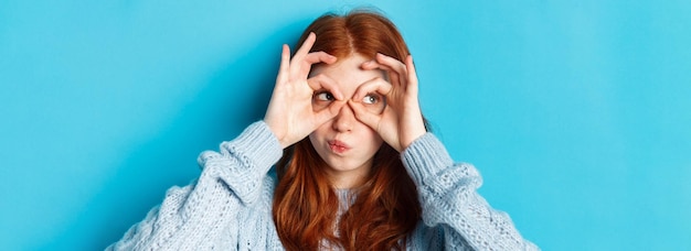 Headshot of funny redhead girl thinking looking through finger glasses with thoughtful face standing