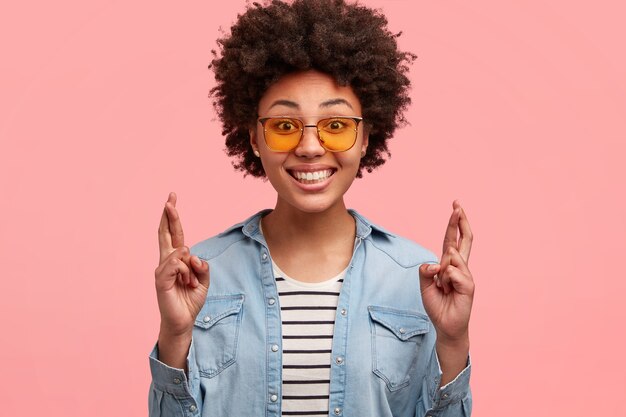 Headshot of glad young Afro American woman eagers dreams come true, crosses fingers for making wish, wears trendy shades, smiles broadly, shows perfect teeth, models against pink wall.