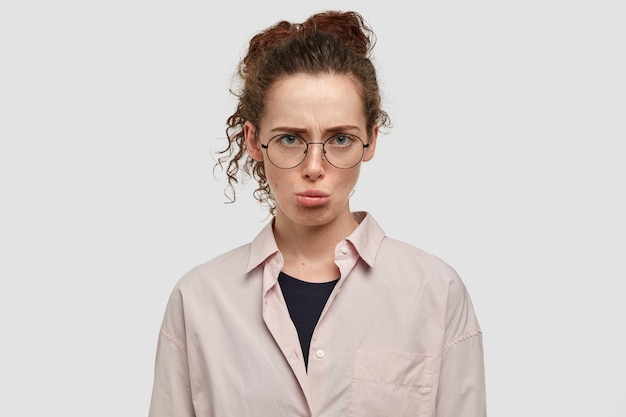 Headshot of gloomy insulted young female feels offended, frowns face, discontent with boyfriend`s bad behaviour, has crisp dark hair, dressed in oversized shirt, isolated over white wall.