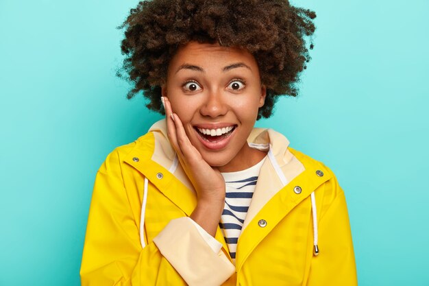 Headshot of good looking African American woman touches cheeks, looks gladfully being in high spirit, dressed in waterproof raincoat, isolated over blue background