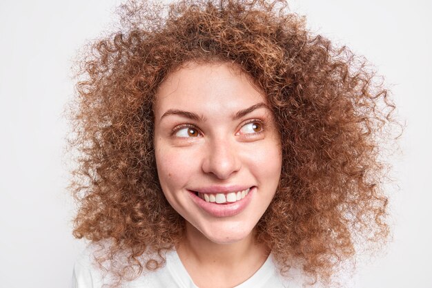 Headshot of good looking cheerful curly girl gazes away in studio against white wall.