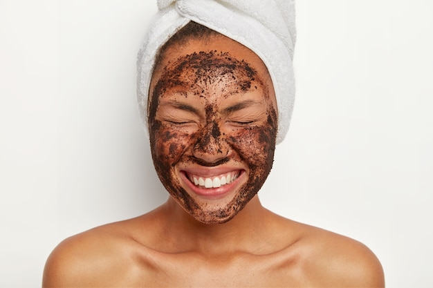 Headshot of good looking overjoyed young woman keeps eyes shut, smiles broadly, shows white perfect teeth, wears wrapped towel on head, peels face with coffee scrub, stands shirtless indoor.