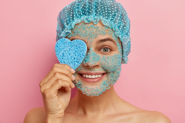 Headshot of good looking pleased woman applies sea salt scrub, holds heart shaped cosmetic sponge, has beauty treatments, naked body skin