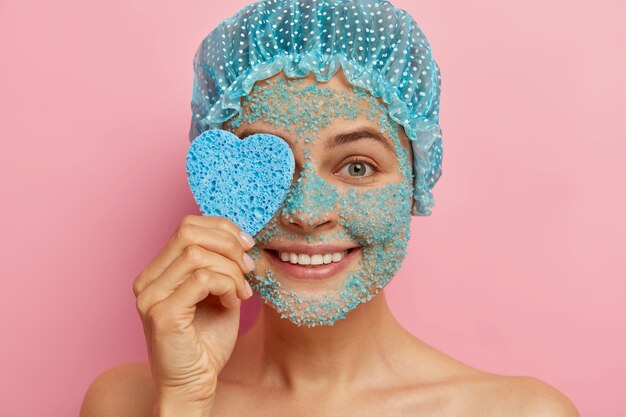 Headshot of good looking pleased woman applies sea salt scrub, holds heart shaped cosmetic sponge, has beauty treatments, naked body skin