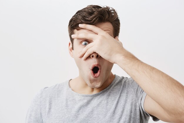 Headshot of goofy surprised bug-eyed young european male student wearing casual grey t-shirt staring  with shocked look, expressing astonishment and shock, hiding face behind his palm.
