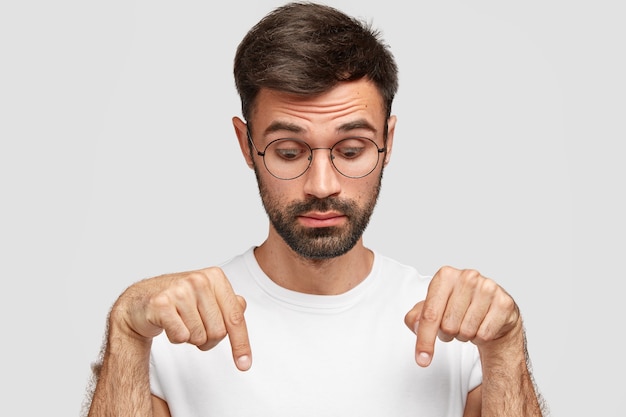 Headshot of handsome bearded male points down with surprised look, notices something on floor, wears glasses, dressed in casual t-shirt, isolated over white wall. People and astonishment