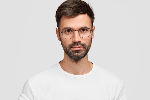 Headshot of handsome male freelancer with appealing look, has dark beard and mustache, looks directly with serious look, wears white casual clothes. Monochrome. Facial expressions.