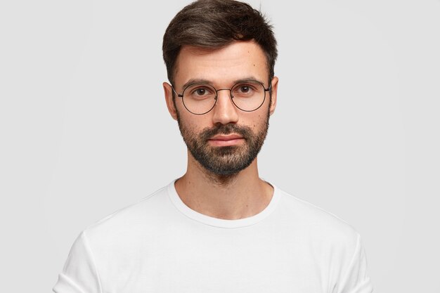 Headshot of handsome male freelancer with appealing look, has dark beard and mustache, looks directly with serious look, wears white casual clothes. Monochrome. Facial expressions.