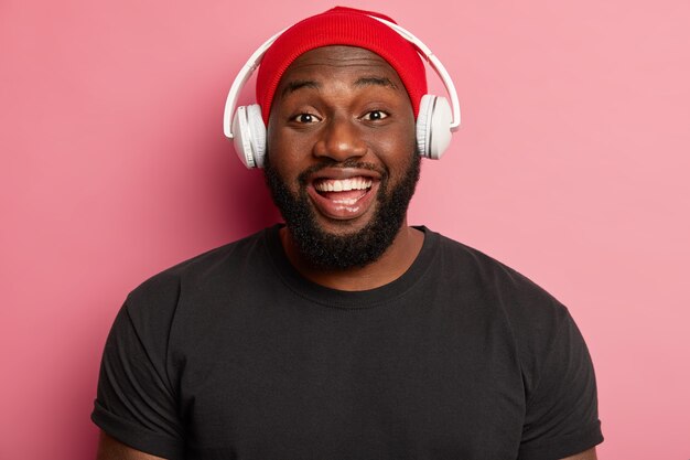 Headshot of handsome man with toothy smile, has white teeth, delivers sound to ears via headphones, listens music in wireless headset