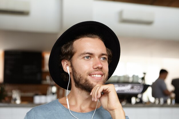 Headshot of handsome young bearded student in black hat smiling joyfully, listening music with earphones