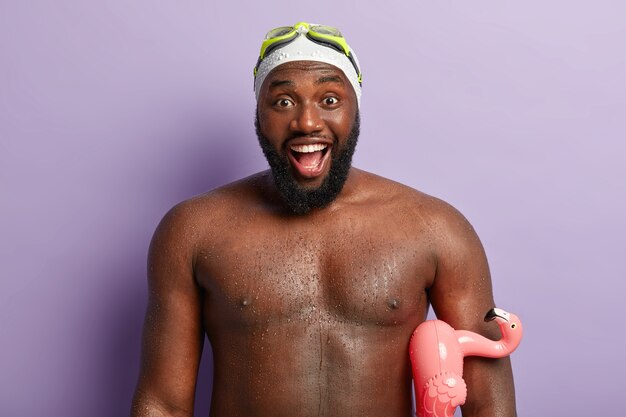 Headshot of happy African American man spends leisure time in resort place, has nice impressions after learning swimming in sea or ocean, holds rubber ring, expresses positive emotions and feelings