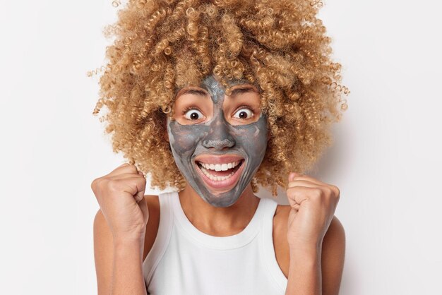 Headshot of happy curly haired woman looks excitedly clenches fists from joy applies facial clay mask for skin treatment wears casual t shirt isolated over white background Skin care concept