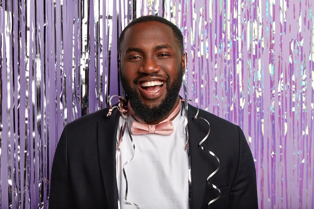 Headshot of happy dark skinned man smiles broadly, shows white shining teeth, dressed in formal outfit, comes on party