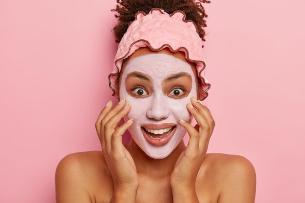 Headshot of happy dark skinned woman applies clay mask, reduces visibility of pores, enjoys effective result of beauty product penetrating deep in skin, stands indoor on pink wall shows bare shoulders