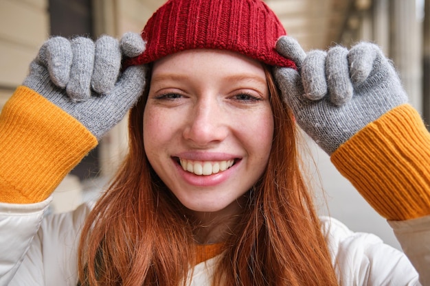 Foto gratuita il colpo alla testa di una ragazza rossa felice con le lentiggini indossa cappello rosso e guanti in inverno cammina per la città