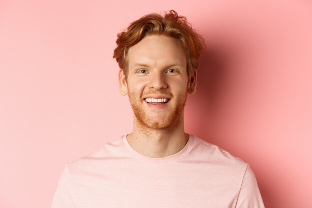 Headshot of happy redhead man with beard and white teeth, smiling excited at camera, standing over pink background.